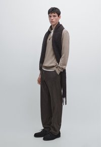 Maglione beige a maniche lunghe con bottoni, sciarpa a frange marrone, pantaloni larghi a righe marroni, scarpe nere, sfondo neutro.
