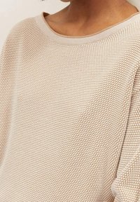 Close-up van een persoon die een beige gebreide trui met textuur draagt, met een ronde halslijn, waarop schouder- en bovenborstgebied zichtbaar zijn.