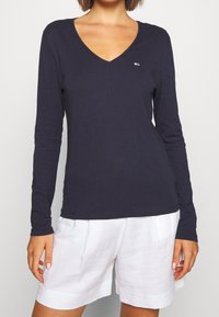 Navy long-sleeve V-neck top made of soft fabric, featuring a small logo badge on the chest and a fitted silhouette. Paired with white shorts.