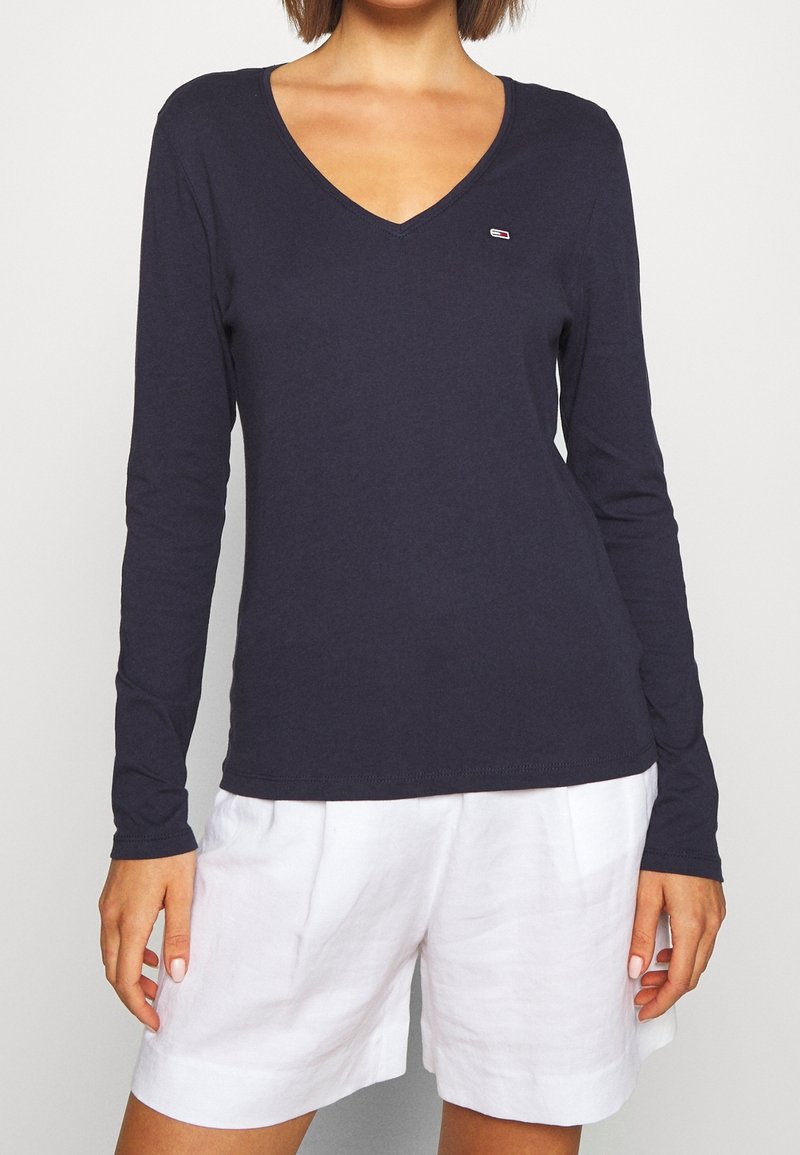 Navy long-sleeve V-neck top made of soft fabric, featuring a small logo badge on the chest and a fitted silhouette. Paired with white shorts.