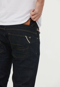 Jeans de mezclilla azul oscuro con costuras amarillas, que cuentan con dos bolsillos traseros y una etiqueta de cuero en la cintura. El bolsillo delantero tiene un detalle de patrón.