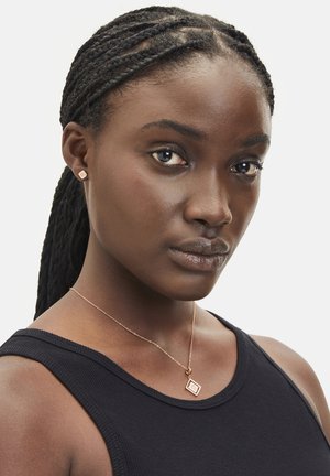 Roséguld halskæde med en firkantet vedhæng, båret af en model. Modellen har flettede hår og har en sort ærmeløs top på.