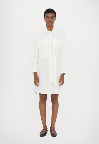 Robe chemise blanche à manches longues, ceinture en tissu, deux poches poitrine, coupe droite et fentes sur les côtés. Portée avec des chaussures à brides noires.