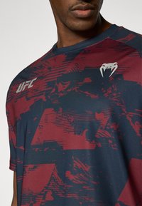 Αθλητικό κοντομάνικο μπλουζάκι σε σκούρο ναυτικό χρώμα με κόκκινο αφηρημένο σχέδιο, που φέρει το λογότυπο UFC και ένα μικρό γραφικό στο στήθος.