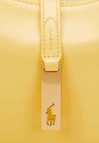 Superficie de cuero amarillo con una etiqueta metálica rectangular dorada que presenta un logo de jugador de polo, sujeta a una correa de cuero cosida.