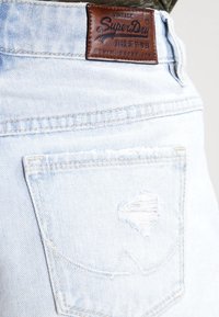 Shorts en denim bleu clair avec une poche distendue visible et une étiquette en cuir brun avec "SuperDry" en lettres en relief.
