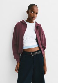 Bordeauxfarbenes Zip-Hoodie mit lockerer Passform, über einem weißen, kurzen T-Shirt mit einem hellblauen Streifen getragen. Marineblaue Hose mit einem schwarzen Gürtel.