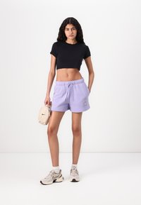 T-shirt noir coupé et shorts lavande avec cordon de serrage. Sac Nike beige et baskets gris clair avec accents. Texture de tissu lisse.