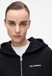 Crna zip-up hoodie od mekog materijala, sa velikom kapuljačom i logom "KARL LAGERFELD" u bijeloj boji na lijevoj strani prsiju.