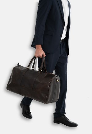 Leather duffel bag in dark brown, featuring sturdy handles, gold hardware accents, and a streamlined shape, carried by a person in formal attire.