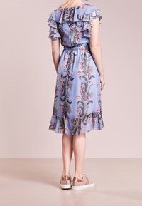 Vaaleansininen kukallinen mekko, jossa on röyhelökaulus ja helma, vyötärö on kiristetty, ja siinä on hienovarainen kiilto, maitopahvin väriset lenkkarit kaverina.