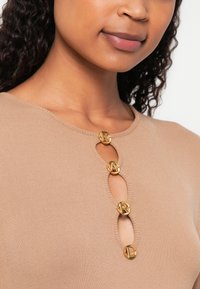 Beige ribbed felső kerek nyakkivágással, négy kör alakú arany gombbal, kiemelt betűkkel és kulcslyuk alakú kivágásokkal az elején.