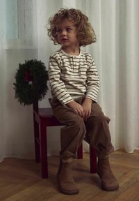 Dziecko w kremowej, pasiasty koszuli z długim rękawem i kieszonką, brązowych spodniach z kordurowej tkaniny i brązowych botkach za kostkę, siedzi na czerwonym krześle z zielonym wieńcem.