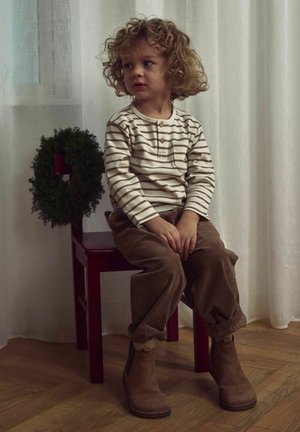 Dziecko w kremowej, pasiasty koszuli z długim rękawem i kieszonką, brązowych spodniach z kordurowej tkaniny i brązowych botkach za kostkę, siedzi na czerwonym krześle z zielonym wieńcem.