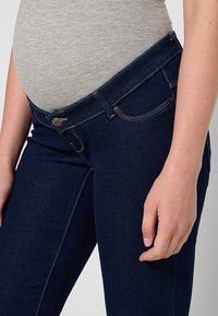 Tumši zili grūtniecības džinsi ar gludu apdari, kurus raksturo zems jostasvieta, kontrastējoši diegi un viens pogas aizvars priekšpusē.