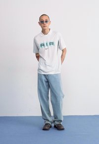Biely polo tričko s grafikou "AIR" v teal farbe, v kombinácii s svetlomodrými voľnými džínami a tmavými teniskami, stojí na modrej podlahe.