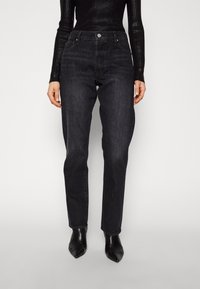 Mustat farkut suoralla leikkauksella, joissa on viisi taskua, metallinappikiinnitys ja hienovarainen kuluminen rakenteen luomiseksi. Yhdistetty kärkipäisiin mustiin saappaisiin.