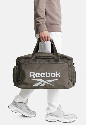 Persona con pantaloni bianchi e scarpe da ginnastica grigie che tiene una borsa da palestra Reebok marrone per i manici al proprio fianco.