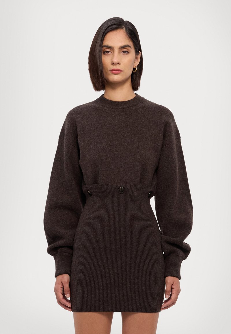 Mørkebrun strikkjole med høj halsudskæring, voluminøse ærmer og ribbet tekstur. Udsmykket med to knapper i taljen.