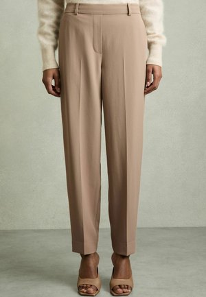Beige, hoch taillierte Hose mit maßgeschneidertem Schnitt, vorderen Falten und ohne sichtbare Taschen. Der Stoff hat eine glatte Textur und wird mit nudefarbenen Sandalen kombiniert.