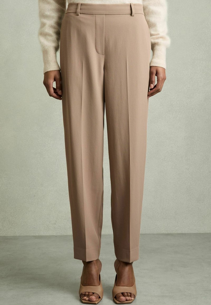 Beige hameidat trouserit, joissa on mukautettu istuvuus, etupleatit ja ei näkyviä taskuja. Kangas on sileäpintainen ja paritettu nude-sandaalien kanssa.