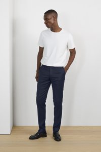 Camiseta blanca de manga corta combinada con pantalones formales azul marino y zapatos negros, de pie sobre un suelo de madera contra un fondo claro.