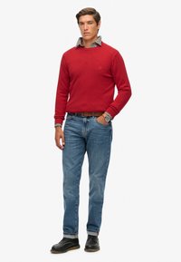Rode trui met een ronde hals, laag over een geruit shirt. Draag het met blauwe jeans en zwarte schoenen, inclusief een bruine riem en horloge.