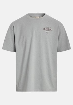 T-shirt gris clair à manches courtes avec petit texte sombre « "GRACIOUS" » sur la poitrine gauche, col rond, design simple, sans personnes.