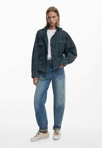 Veste en jean bleu foncé avec grandes poches et capuche à cordon, associée à un jean bleu clair coupe décontractée et des baskets blanches.