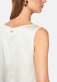 White sleeveless top with a smooth, shiny texture, featuring a round neck and a small silver logo detail at the back. Black statement earrings.