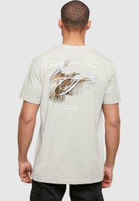 Hellgraues Baumwoll-T-Shirt mit einem detaillierten Vogelgrafik auf der Rückseite und dem Text "Glaube über Angst" in stilisierter Schrift.