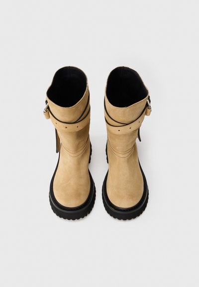 Para brązowych zamszowych botków do połowy łydki z czarnymi grubymi podeszwami i owijającymi paskami, widocznych z góry na białym tle.