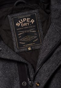 Gewatteerd zwart jack met een grijze, gestructureerde stof, gelabeld "Super Dry", met goudkleurige accentstiksels en een ritssluiting. Maat M.