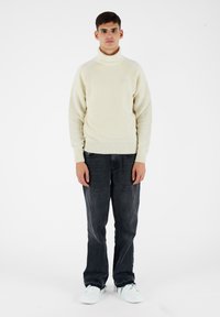 Maglione a collo alto di colore crema chiaro con polsini e orlo a costine, abbinato a jeans grigio scuro e sneakers bianche. Tessuto a maglia liscia, vestibilità casual.
