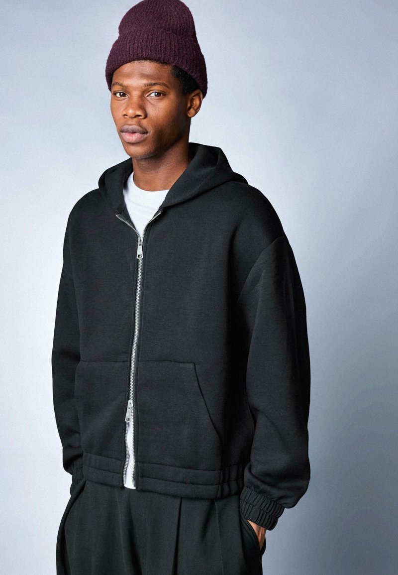 Giovane uomo con cappello di lana bordeaux, felpa nera con zip sopra una maglietta bianca, e pantaloni neri, in piedi contro uno sfondo grigio chiaro.