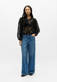 Sort bluse i blonder med lange, puffede ærmer og rund halsudskæring, kombineret med bredbenede blå denimjeans og sorte sko.