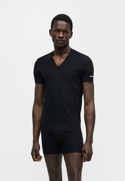 Άντρας που φοράει μαύρο T-shirt με V λαιμόκοψη και μαύρα μποξέρ, στέκεται μπροστά σε απλό φωτεινό φόντο.