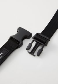 Ceinture en nylon noire avec deux boucles en plastique, une à clip et une glissière. Surface texturée avec une étiquette logo sur la sangle.