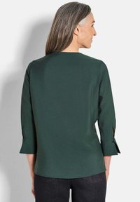 Frau mit langen grauen Haaren, die eine dunkelgrüne Bluse mit langen Ärmeln und schwarze Jeans trägt, von hinten vor einem weißen Hintergrund gezeigt.