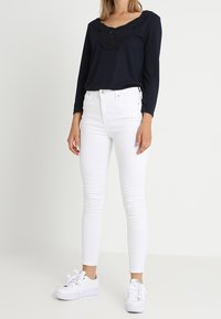 Navy-langärmliges Oberteil mit Rüschenkragen, kombiniert mit eng anliegenden weißen Jeans und weißen Sneakers mit dickem Sohlen und Schleifen-Schnürsenkeln.