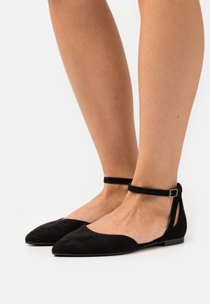 Ballerines en daim noir avec un bout pointu, une sangle à la cheville avec boucle en argent et des découpes sur les côtés. Semelle plate et texture lisse.