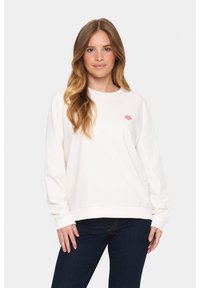 Felpa beige chiaro a maniche lunghe, con polsini e orlo a coste, che presenta una piccola stampa di labbra rosa sul petto. Indossata con jeans scuri.