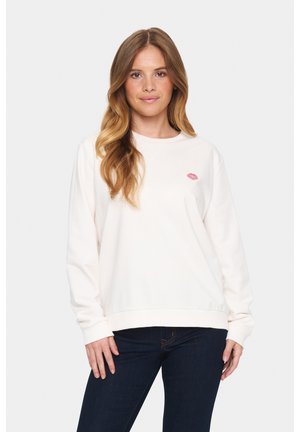 Lichtbeige sweatshirt met lange mouwen, geribbelde manchetten en zoom, met een kleine roze lippenafdruk op de borst. Draagt het met donkere spijkerbroek.