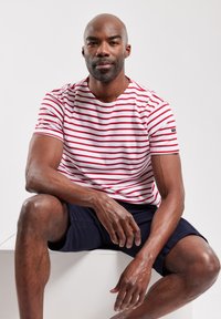 Camiseta de cuello redondo a rayas rojas y blancas, de mangas cortas, combinada con pantalones cortos azul marino. El modelo se sienta en un pedestal blanco, mostrando un look casual.