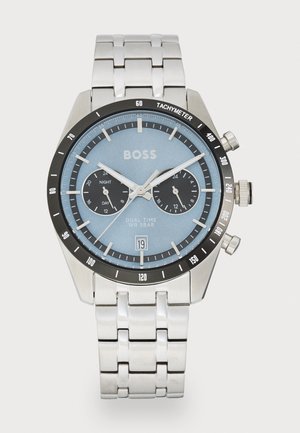 Rustfritt stålklokke med blå urskive, svart tachymeter bezel, doble tidsunderurskiver og sølvfarget armbånd. Har et datovindu.