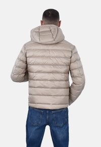 Giacca imbottita beige con cuciture orizzontali e cappuccio, caratterizzata da un esterno liscio e lucido e polsini elasticizzati, abbinata a jeans blu.