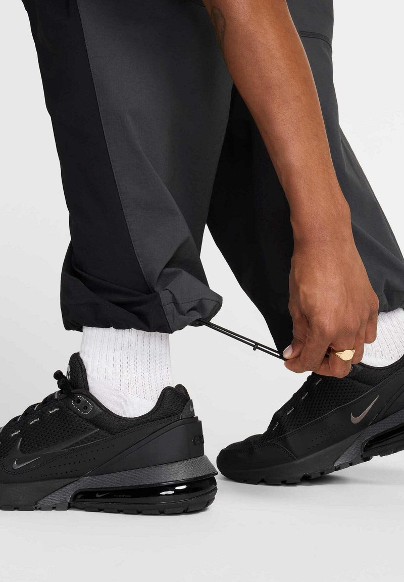 Zapatillas deportivas negras con parte superior de malla texturizada y acentos sintéticos, combinadas con pantalones negros con puños elásticos y calcetines blancos de caña media.