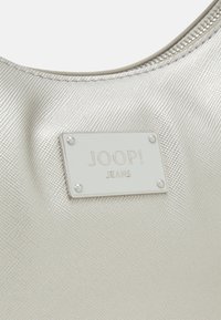 Sac à main en tissu texturé or clair avec une plaque rectangulaire en argent gravée "JOOP! JEANS" fixée par quatre rivets.