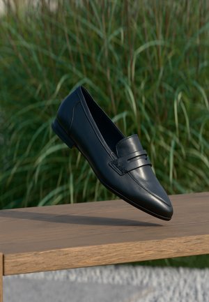 Zwarte leren loafer zweeft boven een houten bank met groen hoog gras op de wazige achtergrond.