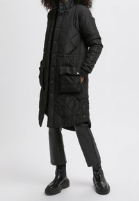 Abrigo largo negro acolchado con cuello alto, botones de presión, bolsillos laterales y un acabado elegante y brillante, combinado con pantalones negros ajustados y botas.